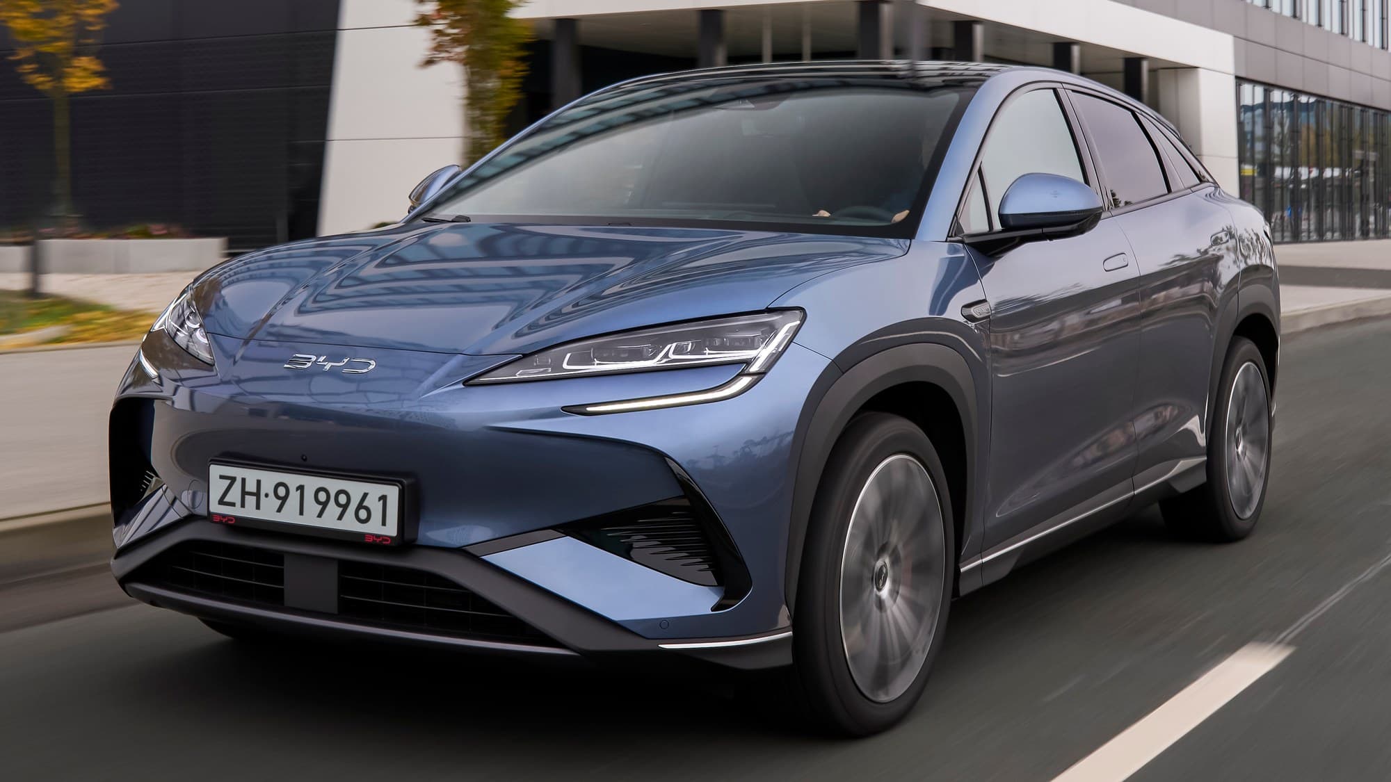 Blue-grey BYD Sealion 7 electric SUV driving on a city street past a modern office building